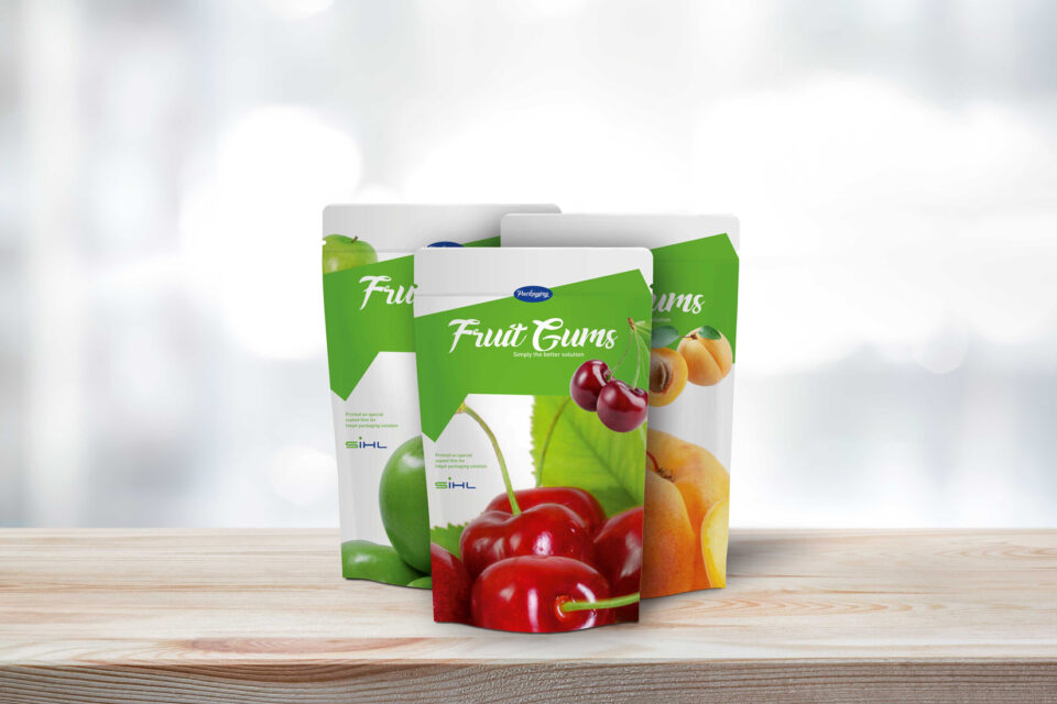 3 Standbodenbeutel für FruitGums mit Kirschen, Äpfeln und Pfirsichen auf Holzplatte vor grauem Hintergrund
